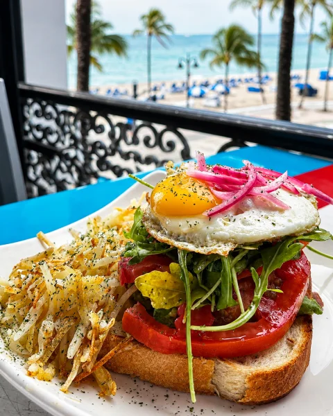 Avocado toast topped with tomato, greens, pickled onions and a fried egg overlooking Fort Lauderdale Beach.