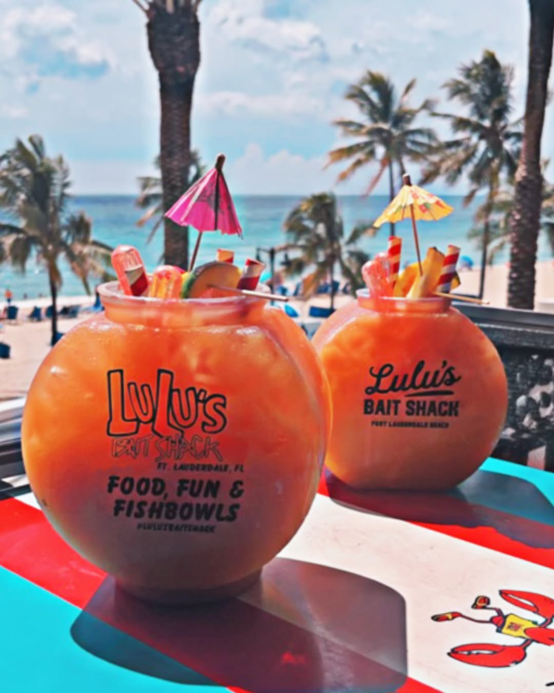 Two Lulu's Bait Shack signature fishbowls on the beachfront patio, palm trees and ocean behind them.